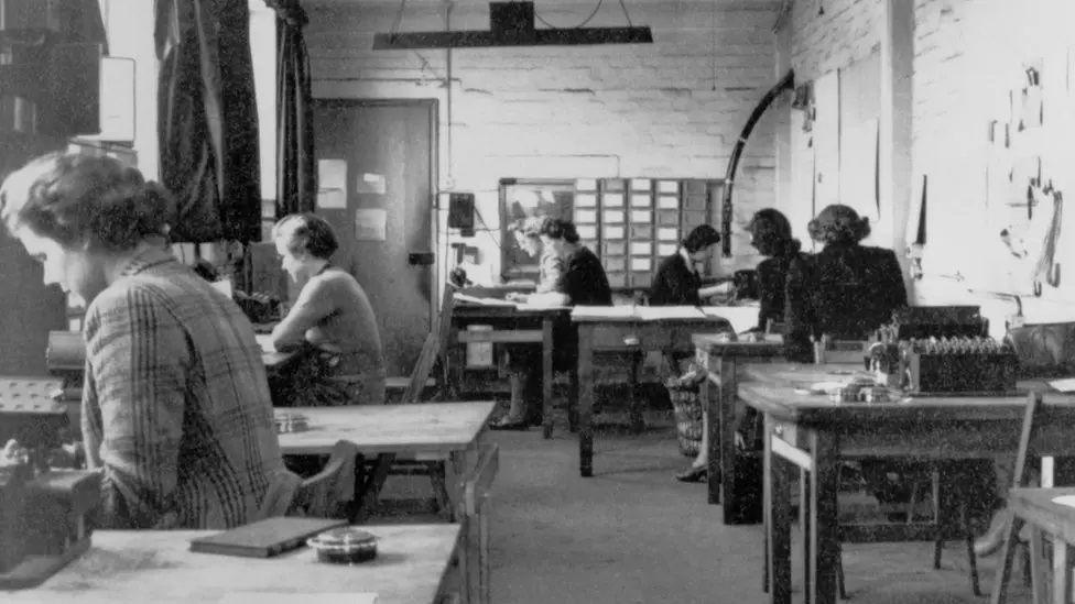 Wooden tables at which women are sitting, working with old-fashioned code-breaking machines with small circular buttons. There are plain white walls, and curtained windows to the left. There is a light hanging from the ceiling.