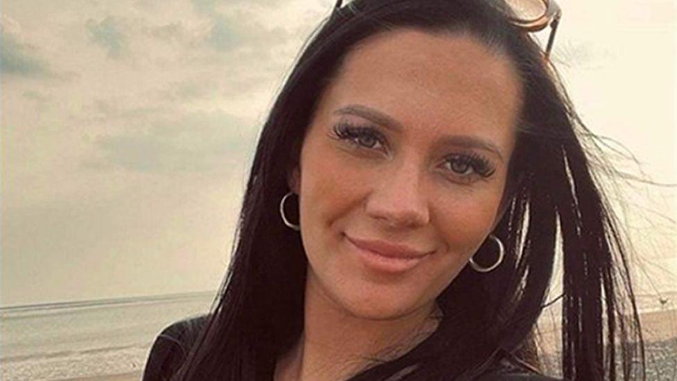 Kiena Dawes, with long dark hair, long eyelashes and silver hoop earrings, smiles at the camera in front of a beach