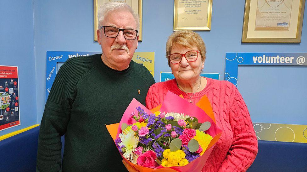 Graham Gedney in dark shirt and glasses stands next to his wife, Linda Gedney, who is wearing a pink jumper and has red glasses and is holding a bouquet of flowers