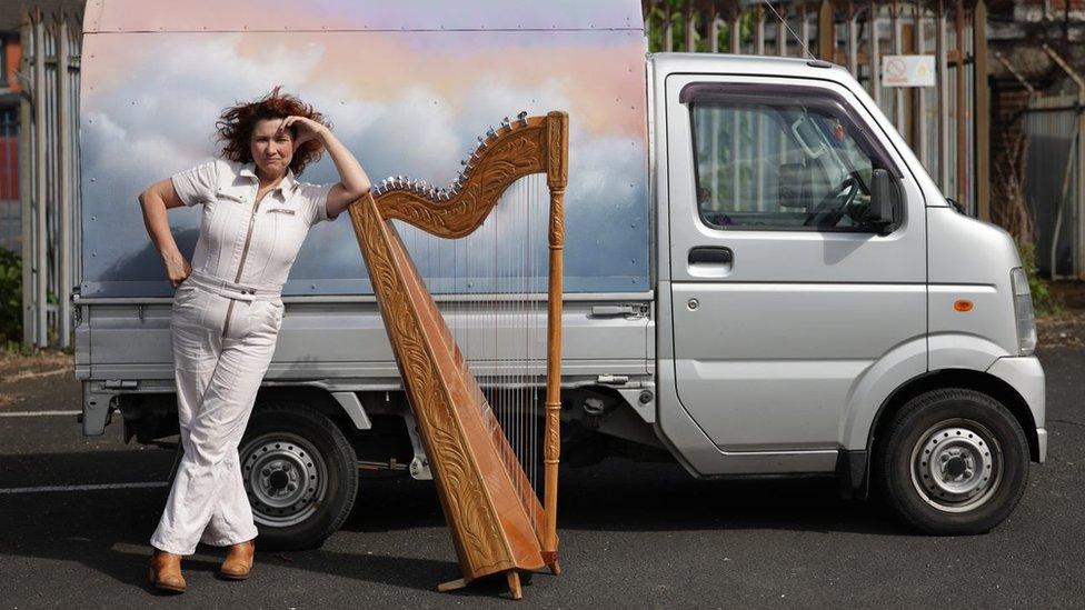 Ursula Burns wearing a white jumpsuit, leaning on her harp with her pastel pink and blue van in the background