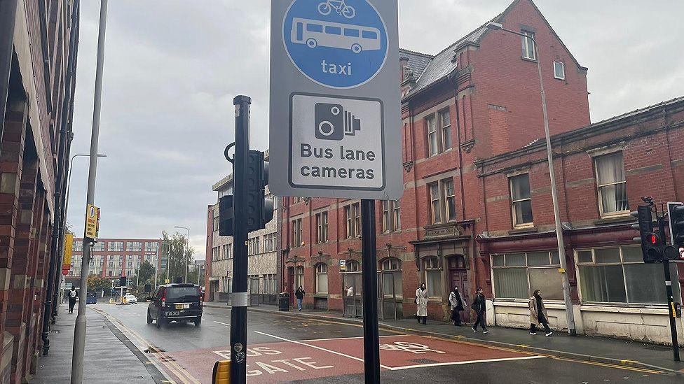 Street scene in Preston showing red brick buildings either side of a road. The road says 'bus gate' in white on a red background on the left hand side, with a picture of a bicycle on the other. A black cab is driving into the distance and a group of people are walking on the far side of the street