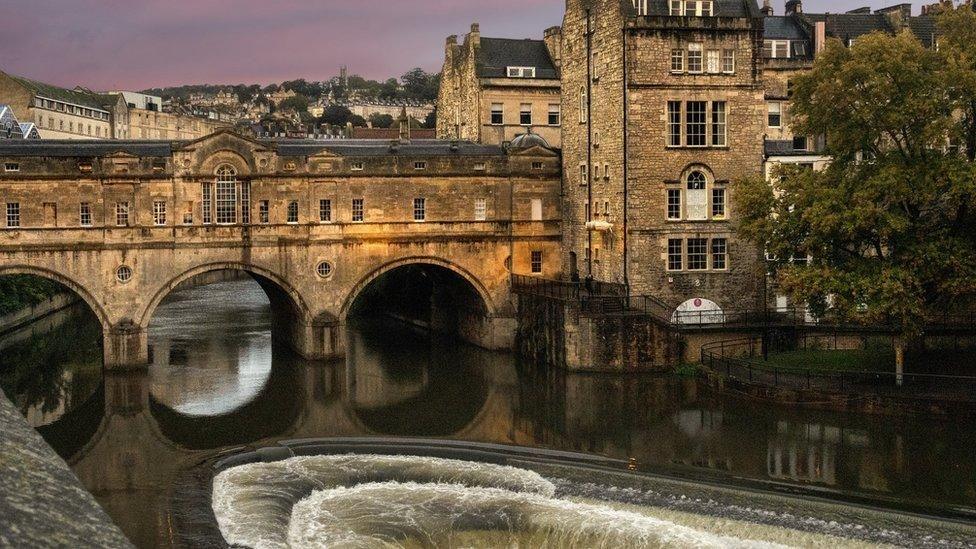 The river flowing down cascading steps in Bath with Georgian buildings in the background and a bridge with three tunnels underneath it. 