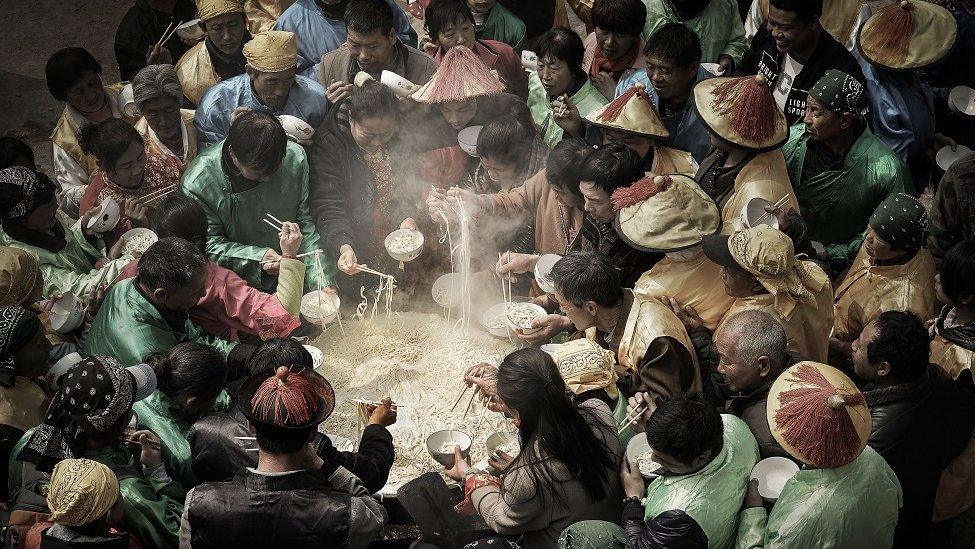 People stand around a giant cauldron eating noodles