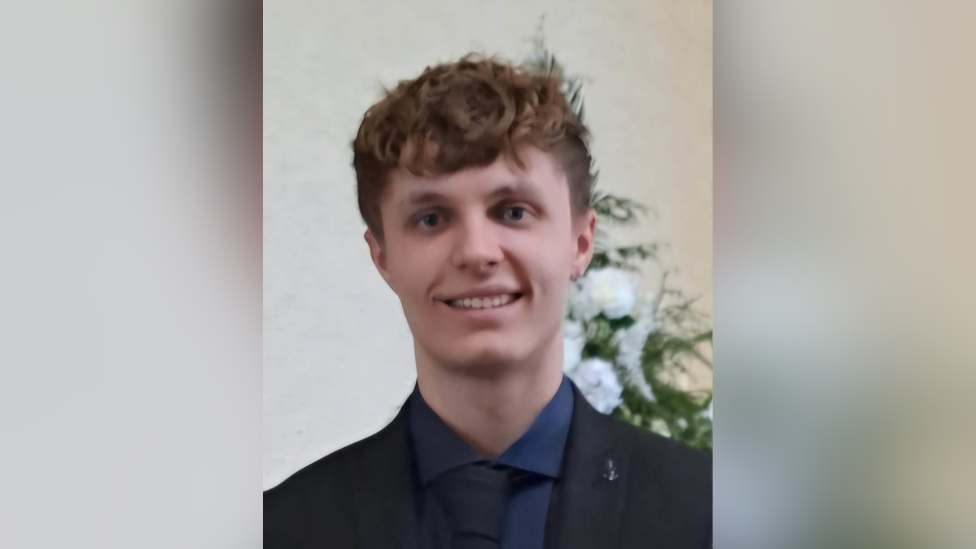 A still image of Kacper Dudek. He is a young man with short, light brown hair and is wearing a black suit jacket, a black tie and a navy shirt. He is standing in front of a bouquet of white flowers.