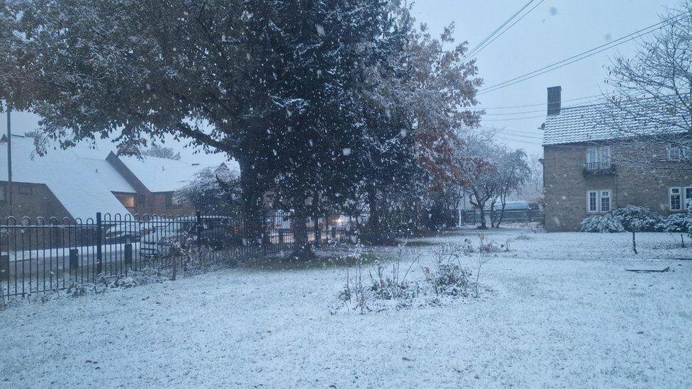 Metal fence around a garden covered in snow with homes seen surrounding - snow if falling