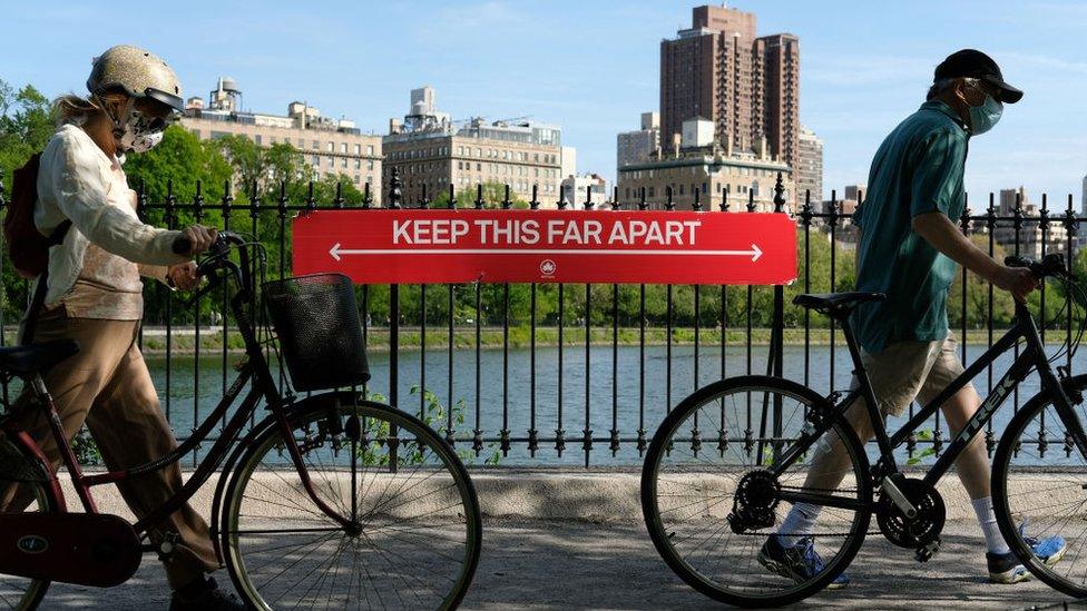 People wearing protective masks walk their bicycles past a social distancing sign reading "KEEP THIS FAR APART" at Jacqueline Kennedy Onassis Reservoir in Central Park during the coronavirus pandemic on May 17, 2020 in New York City