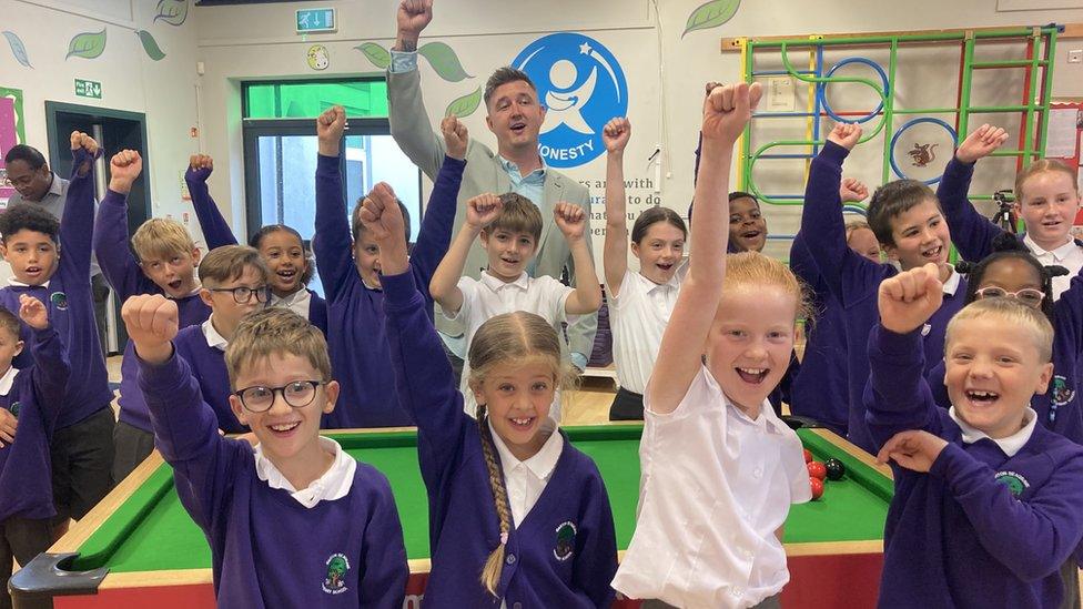 Children, mostly wearing blue school sweaters, and Kyren Wilson stand around a snooker table with their right arms up