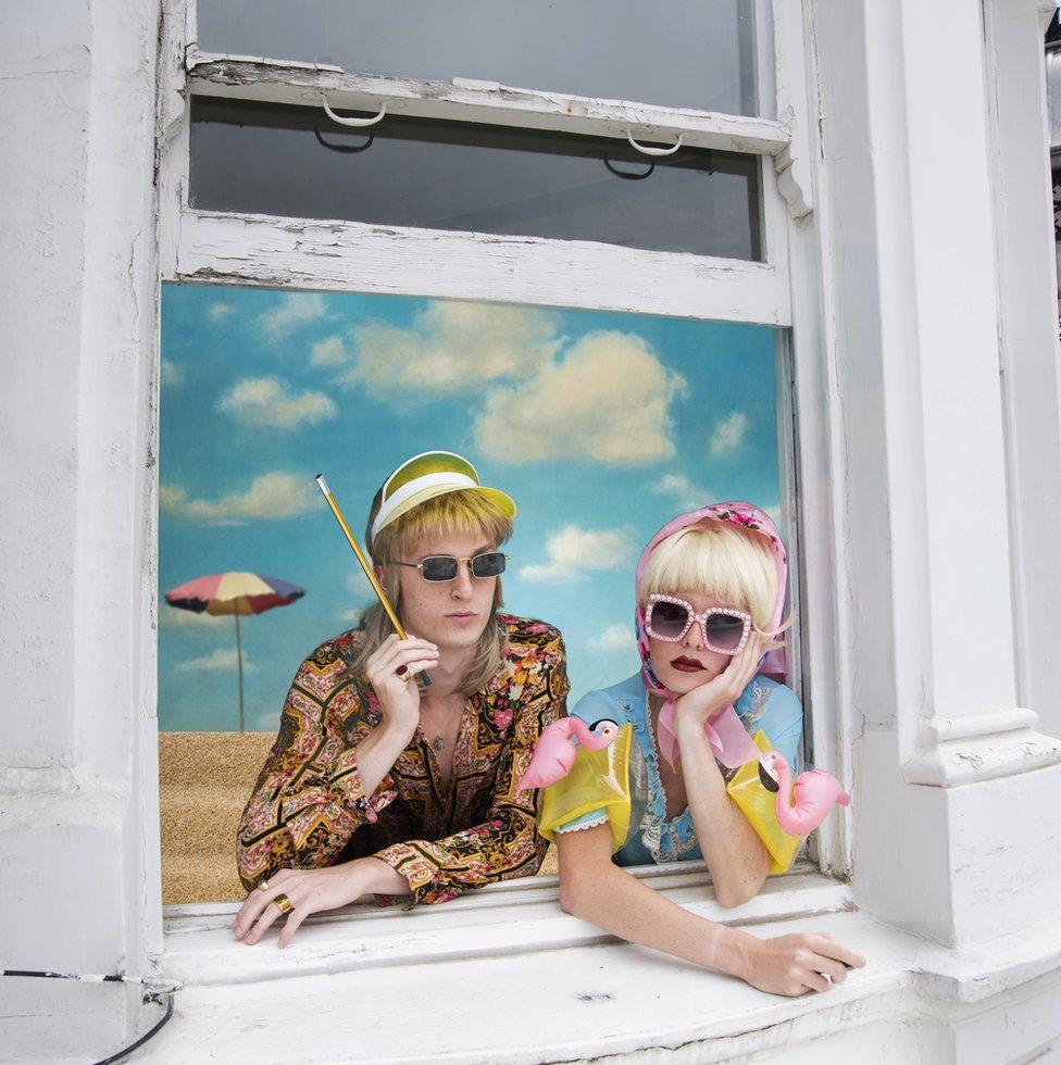 A couple dressed in colourful summer clothes look out of a window on to a beach