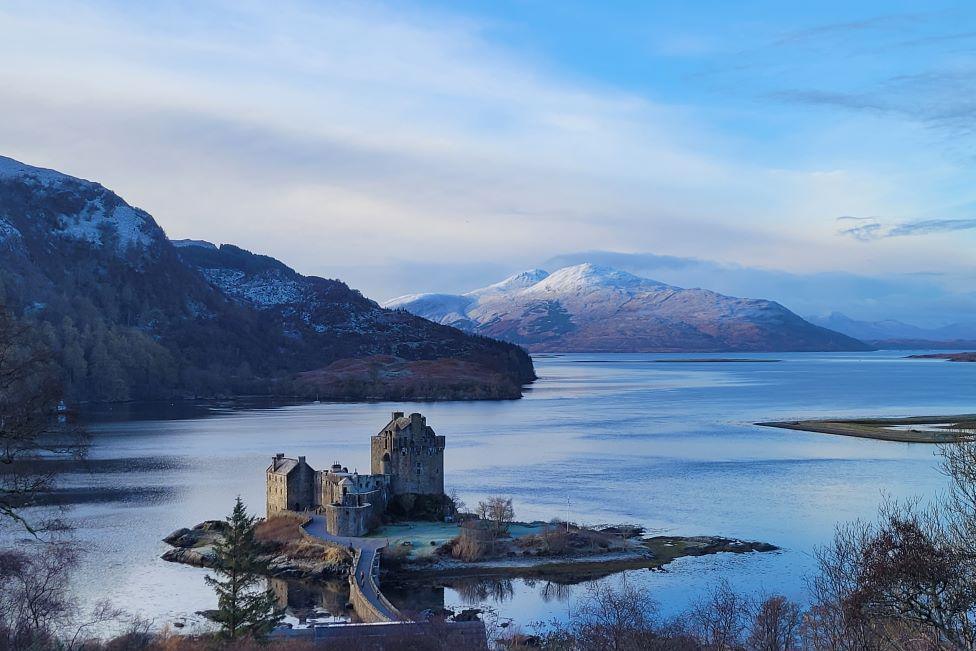 Castle on small island in lake, with frosty snow-covered mountains in the background, image framed by tree branches.