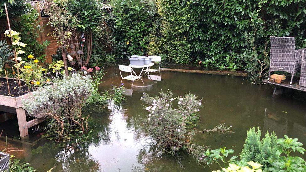 A garden with many bushes and plants filled with floodwater. In the background two patio sets are half submerged.