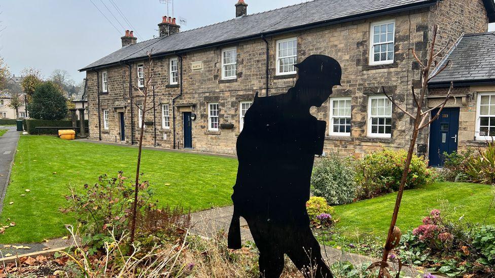 General view of the village showing a row of stone cottages behind a rectangular patch of neat grass. There is a black silhouette of a soldier in the foreground