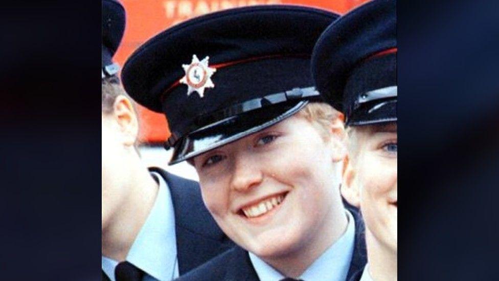 Fleur Lombard, pictured in uniform. She is standing with other people also in uniform but the picture is cropped so you can only see her face fully. She wears a large cap with the Avon Fire and Rescue emblem on it, and a light blue shirt, dark tie and dark blazer. 