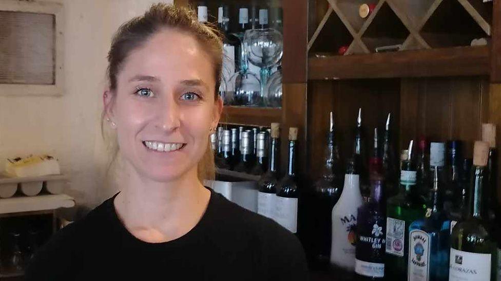 Silvia Panés Cañadas smiling and standing in a bar area of a restaurant with bottles of wine and spirits lined up behind her