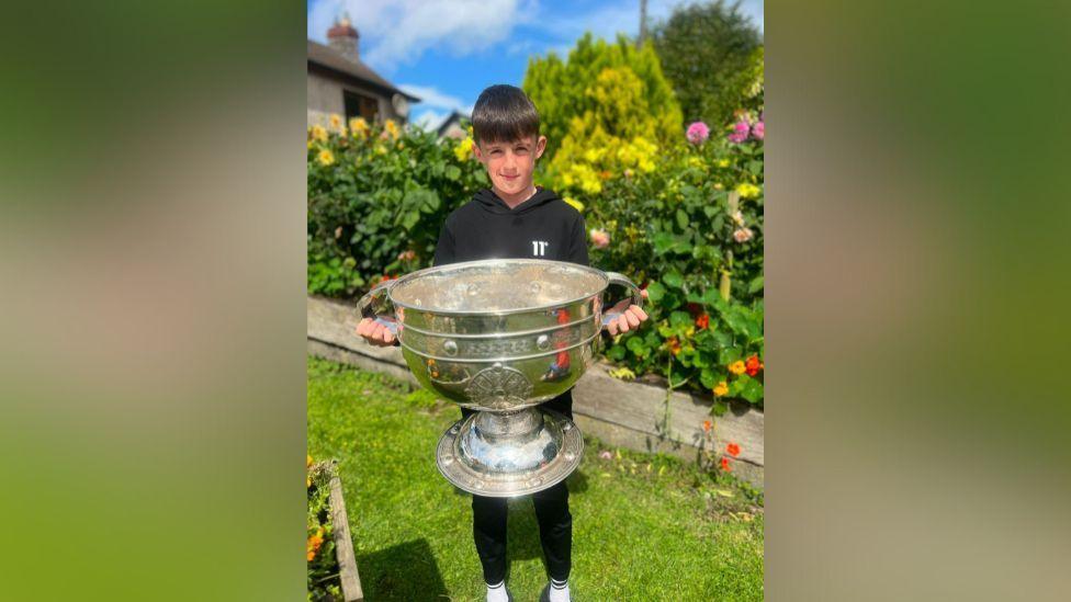 Tiarnán Trainor wearing a black hoody, holding the All Ireland GAA football trophy. He is standing on grass, with a flower bed behind him.