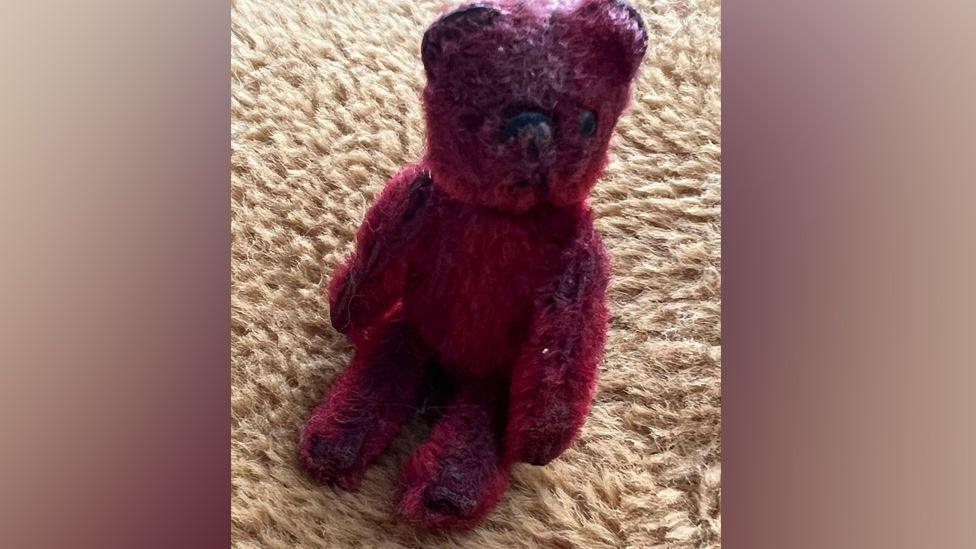 A tatty red teddy bear sitting on a carpet