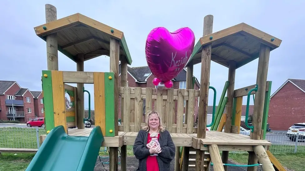 Ms Flux-Edmonds in front of a wooden play area, with a slide to the left of the picture and a climbing area to the right. She is standing and holding a soft toy, and above her is a pink balloon that says "Keziah".