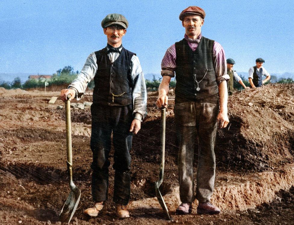 Two men stood with digging tools and soil and walls behind