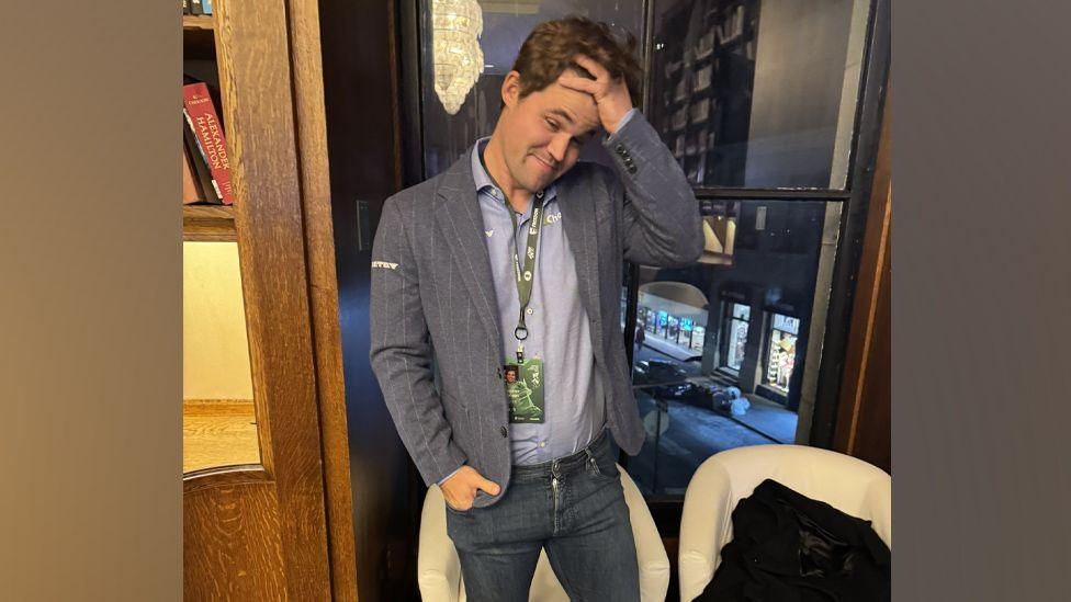 Magnus Carlsen, in a shirt, grey suit jacket and grey jeans, runs his hand through his hair