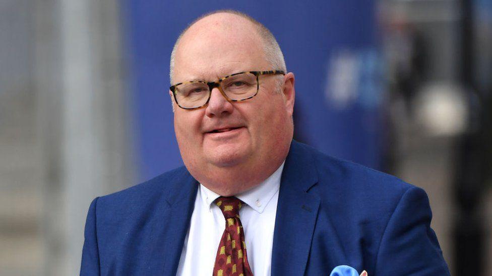 Lord Pickles, who is wearing glasses, a navy suit with a white shirt, and a red and yellow tie.