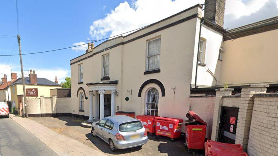 The Park Hotel viewed from the right side. It is a small two-storey hotel that is built straight onto the road, with a pavement in between. It is white and has five red Biffa bins outside the front, and a silver car parked on the forecourt. The entrance porch has four pillars. There are three sash windows upstairs and two arched windows downstairs.