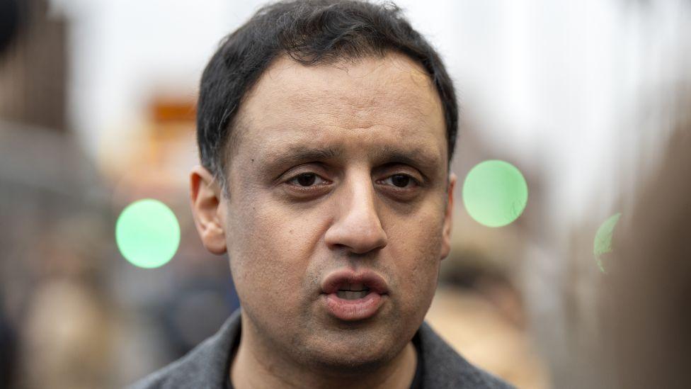 Anas Sarwar, with dark hair and wearing a grey jacket, looks towards the camera while standing outside with blurred traffic lights in the background 