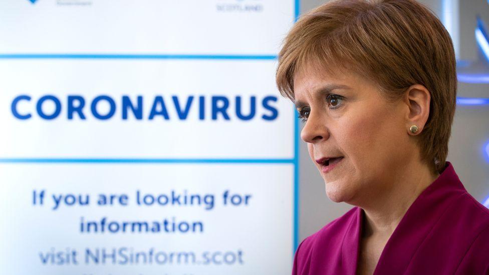 Nicola Sturgeon, with light brown hair and wearing a purple top, stands to the right of shot in front of a coronavirus sign 