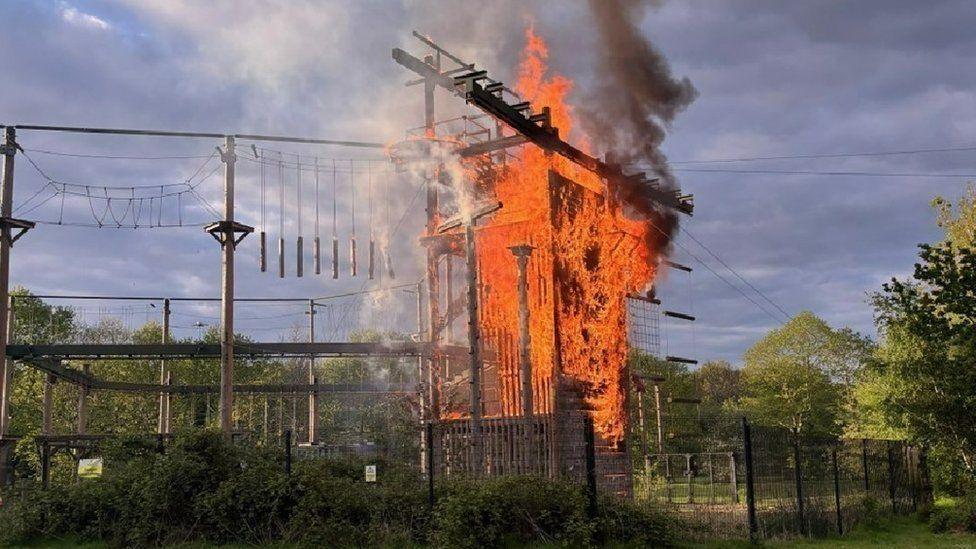 Fencing around a high obstacle course with various structures, wires and netting - a large platform area is engulfed by fire.