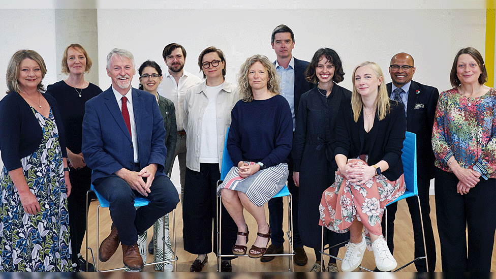 12 people - eight women and four men stood/seated in a semi-circle for a photo-opp.