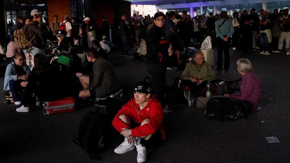 A man sits on the ground in a crowd of other people sitting with their luggage, in darkness outside a train station.