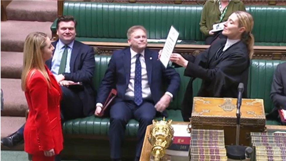 Woman in read reads a card held up by an official in the House of Commons chamber