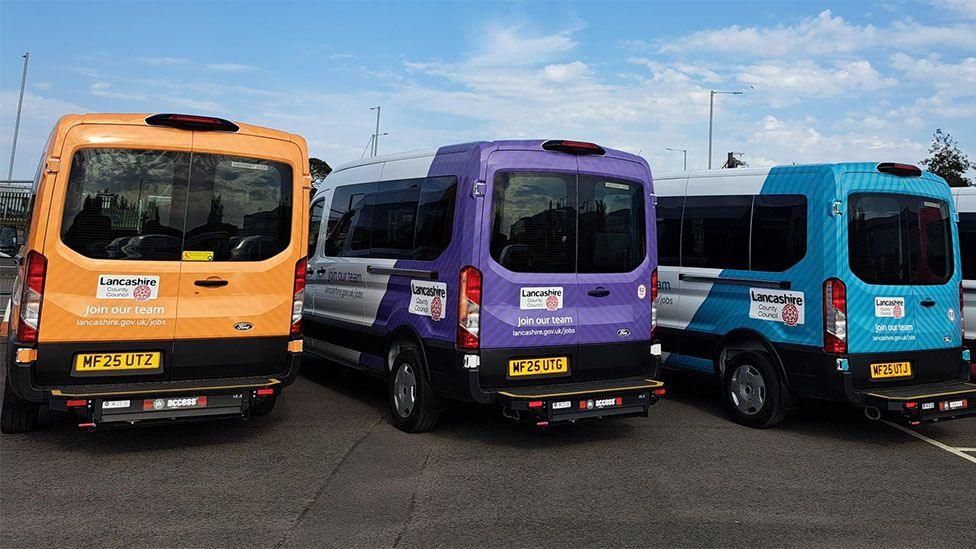 The backs of three of the minibuses parked up on a car park under a blue sky, one is orange, the middle one purple and the one on the right is turquoise