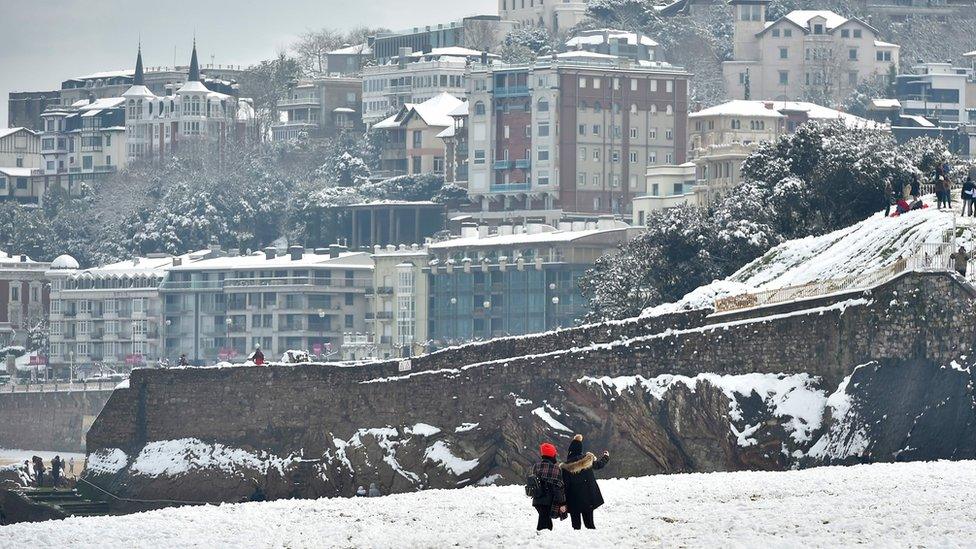 Two people take photos from a white-covered beach with the seaside resort town behind them