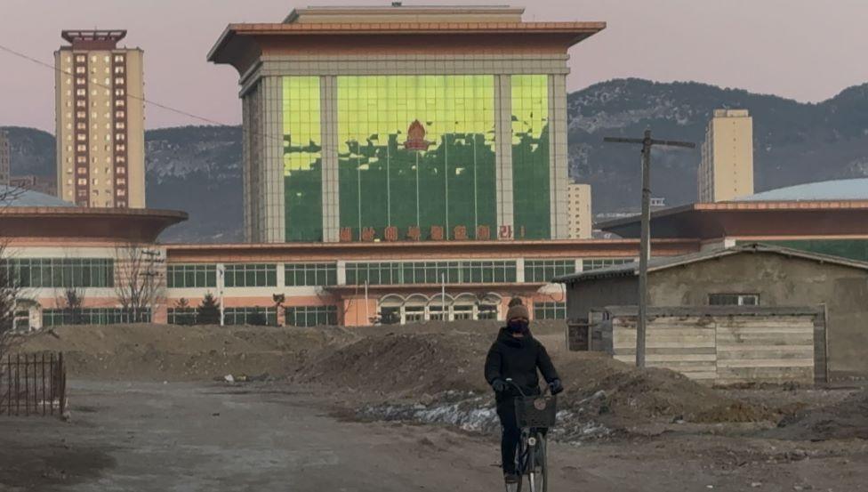 A person dressed in black is cycling on a bike in front of a large building and piles of rubbles. There are tree-topped hills in the background