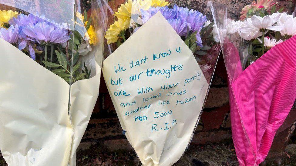 Three bouquets of flowers which have been placed against a brick wall. One bouquet has bright pink wrapping and the other two (in the centre and on the left) are wrapped in a cream colour paper. Written on the bouquet in the centre a message reads: "We didn't know you but our thoughts are with your family and loved ones. Another life taken too soon. RIP."