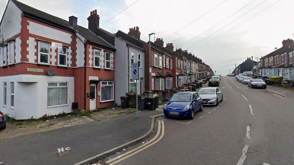 A terraced street in Althorpe Road, Luton, rising up into the distance. Cars are parked on either side of the street. The street sign Althorpe Road is on the first house on the left. Telegraph wires are running down to several houses. 