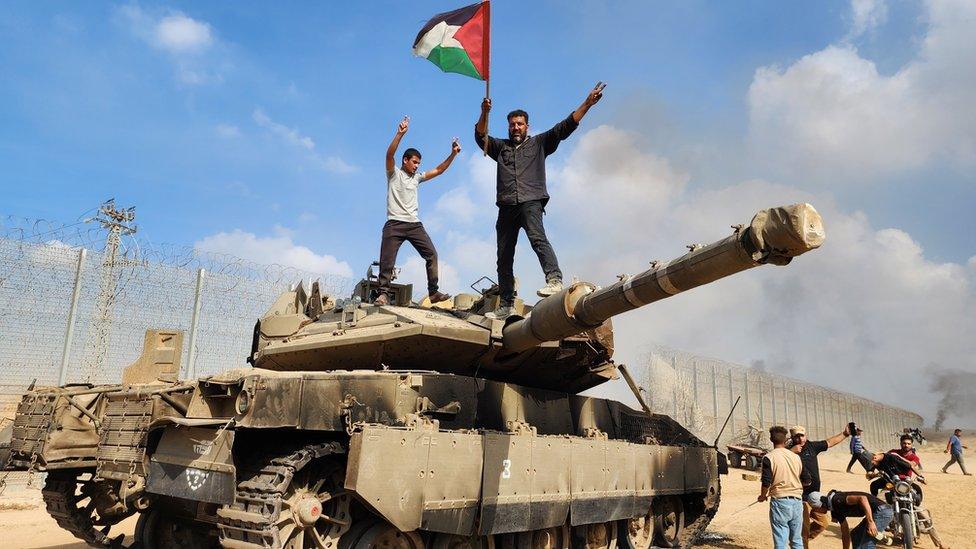 A person waving a Palestinian flag on top of a captured Israeli tank on 7 October 2023