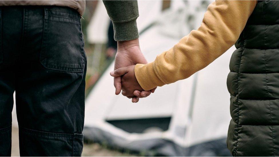 Rear view of unrecognizable parent of elder brother holding hand of child against tent in migrant camp