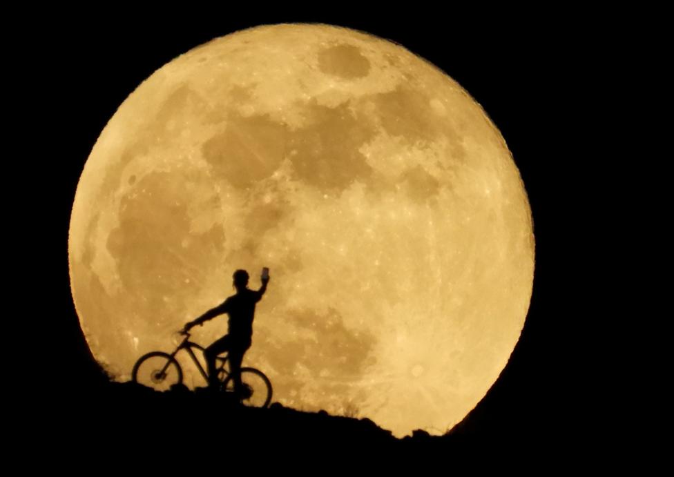 A cyclist uses a cellphone to photograph the full moon, known as the "Super Flower Moon" as it rises over Arguineguin, in the south of Gran Canaria, Spain. 26 May 2021.