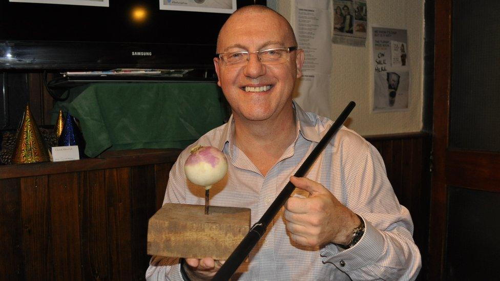 A man smiles while he holds a turnip trophy and a black pole.