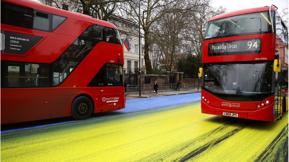 Two buses drive through the yellow and blue paint left by protestors outside the Russian embassy