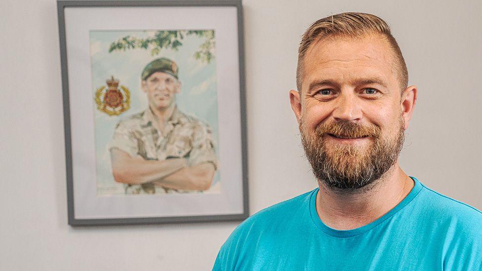 Andrew Powell, of Healthier Heroes, has light brown hair and a beard. He is wearing a bright blue t-shirt as he smiles while standing in front of a franed picture in the wall of a soldier.