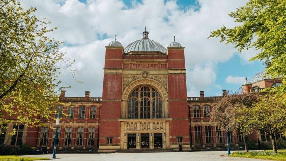 A grand red brick building, topped with cupolas. The building stands with a paved walkway leading to it, between green-leaved trees.