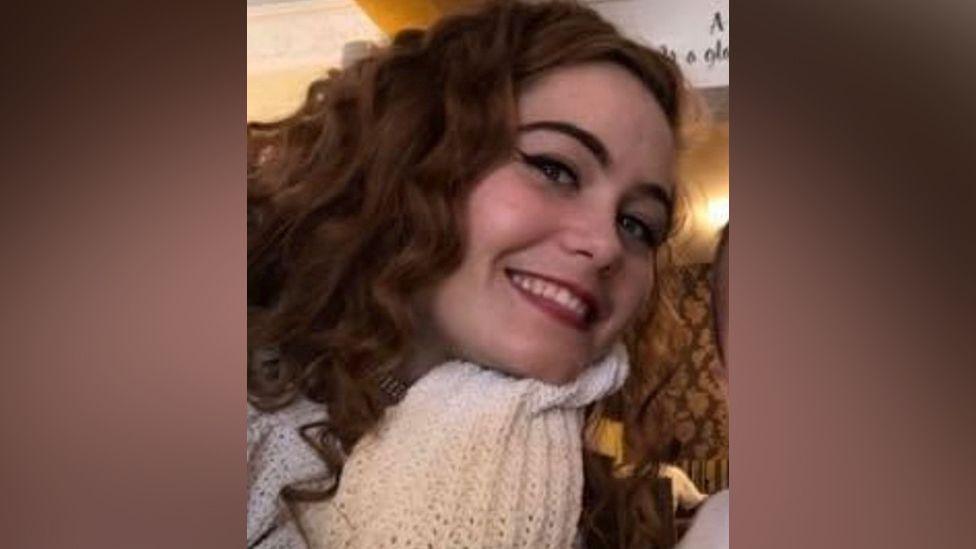 A smiling Courtney Mitchell looks directly at the camera with her head tilted to her left. She is a young woman with long, slightly curly dark brown/ginger hair and is wearing a white scarf. 