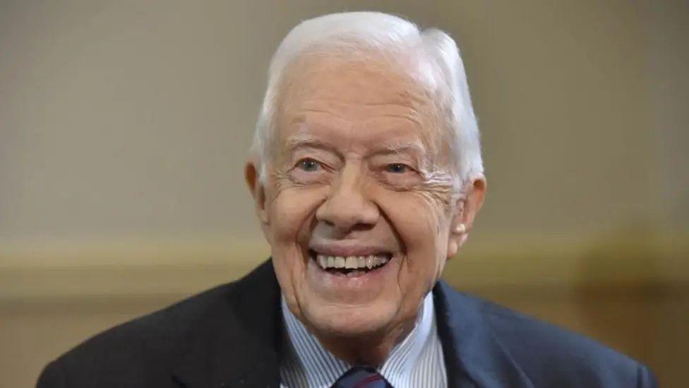 Jimmy Carter is pictured laughing with a broad smile looking slightly beyond the camera, aged 92. He is wearing a smart suit jacket and striped shirt with a dark red tie