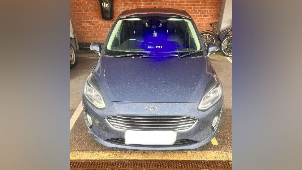 A picture of the car issued by the police. It shows a silver/grey Ford parked in what appears to be a covered parking space. The blue light is clearly visible flashing in the front seat. 