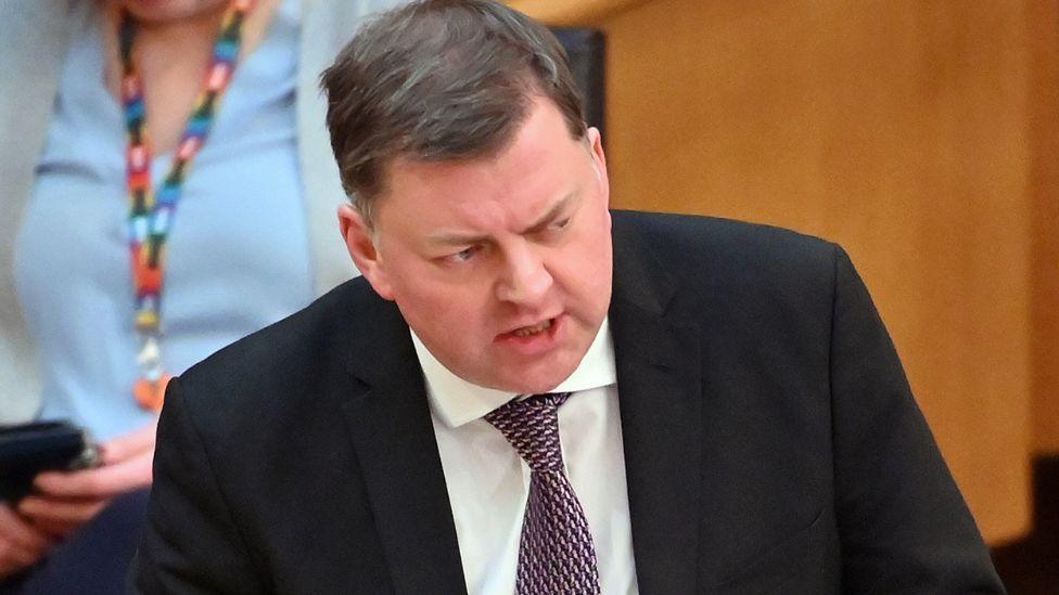 Colin Smyth, a man with dark hair, speaking in the Scottish Parliament. He is wearing a dark suit, white shirt and purple tie. There is wooden panelling and a person using a phone in the background.
