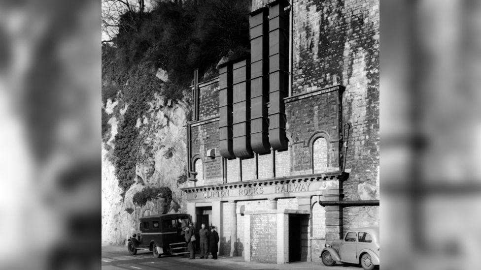 A black and white image of the Clifton Rocks Railway exterior entrance. It is a brick tunnel carved into the cliff, with four metal ventilation shafts pointing upwards. There are two cars parked outside the entrance. 