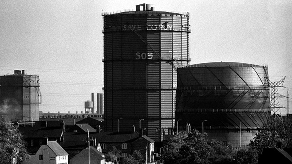 Large circular chimney with smaller gasometer alongside and another chimney in the background. There are houses dwarfed by the structures in the foreground.