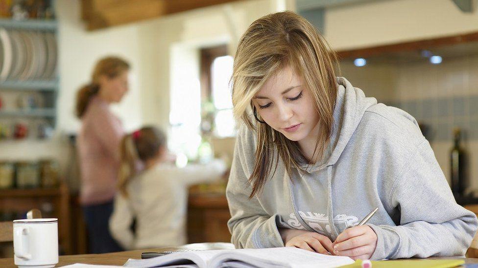 A student revises at home with a book open in front of her. She is wearing a grey hoodie and appears to be working at a kitchen table, with her mum and younger sibling stood at a sink in the blurred background.