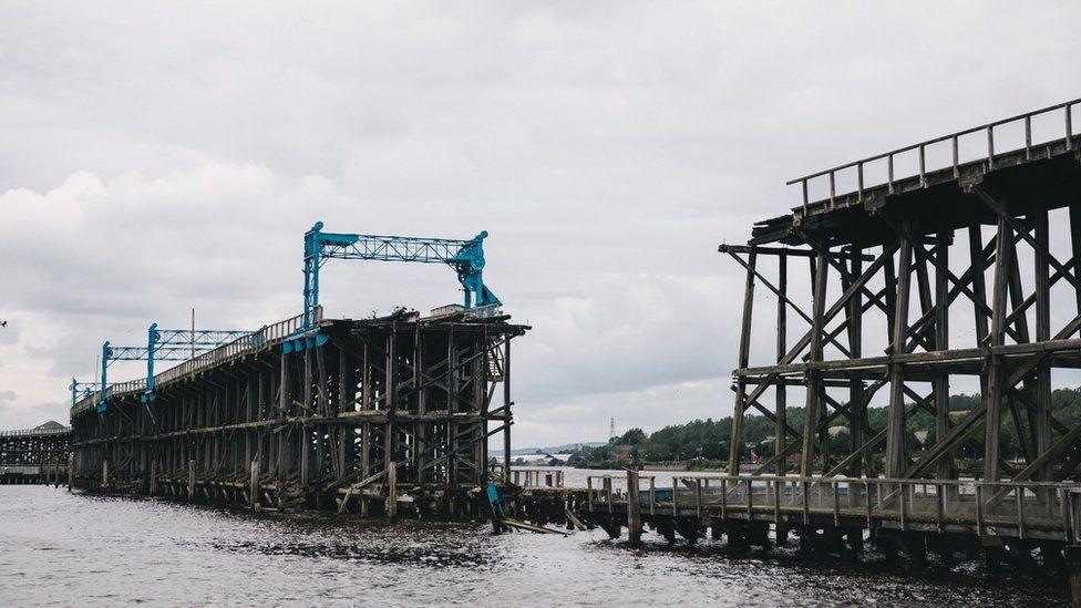 A wooden structure on the River Tyne, at Dunston. A large section in the middle is missing having been destroyed by a fire some years ago. Several metal support structures, painted blue, are in place on the top deck. The river is in the foreground with a cloudy sky above.